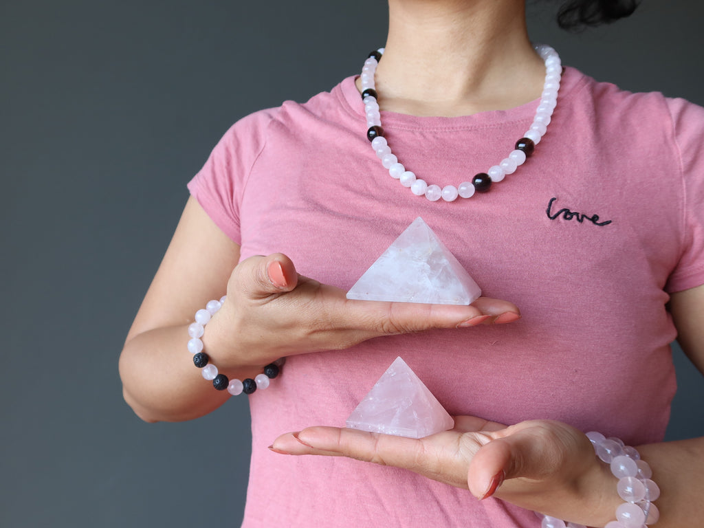 female in pink shirt wearing rose quartz jewelry and holding two pyramids