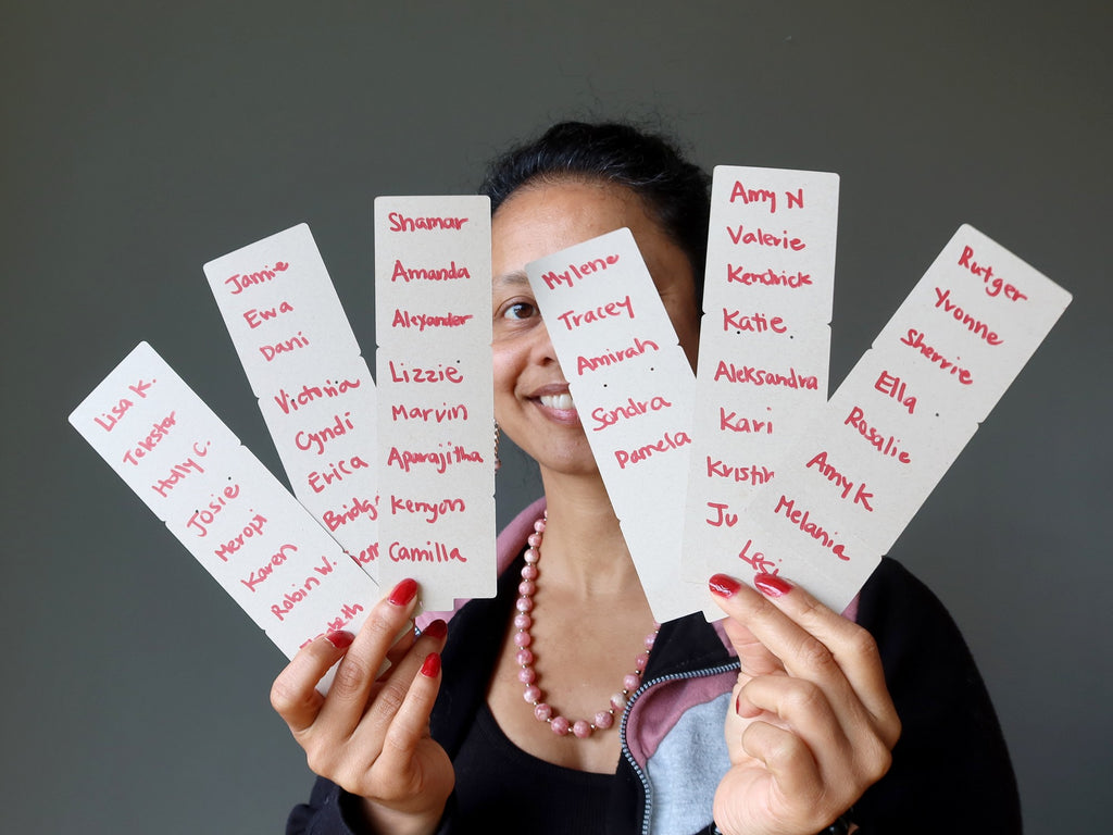 sheila of satin crystals holding up 6 strips of paper with contestant names written down