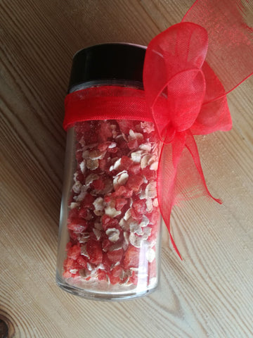 A small glass jar lying on a wooden table. It contains dyed red salt crystals and oats. There is a red ribbon tied around the jar.