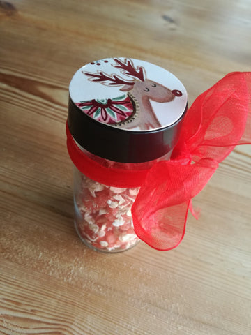 A small glass jar containing dyed red salt crystals and oats. There is a red ribbon tied around the jar and a picture of a reindeer on the lid.