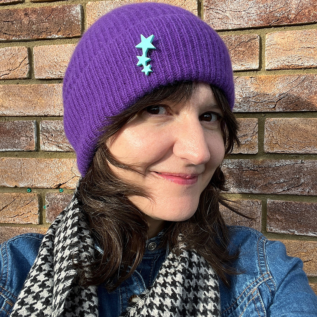 A woman with dark hair wears a purple woolly hat with a tiny star shaped pin attached to it.