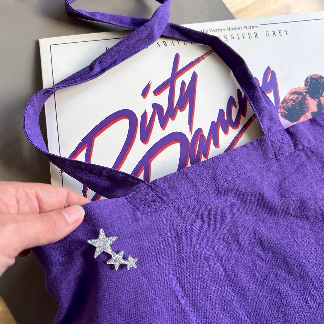 A purple tote bag with a tiny glittery star shaped pin attached. In the bag we can just see a vinyl record.