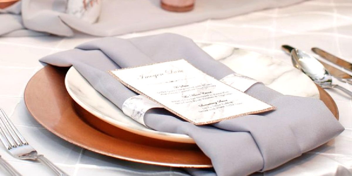 Table setting with chargers: Brown charger, white plate, grey napkin, menu card, silverware on textured tablecloth