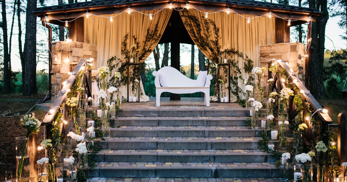 Outdoor wedding with white bench, flowers, and best lights for party