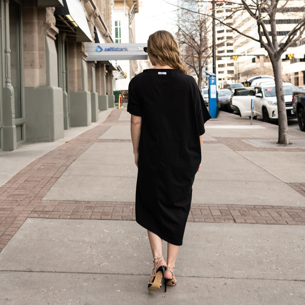 A woman wearing a long black asymmetrical shift dress in organic cotton with asymmetrical sleeves a true sustainable dress by Malaika New York