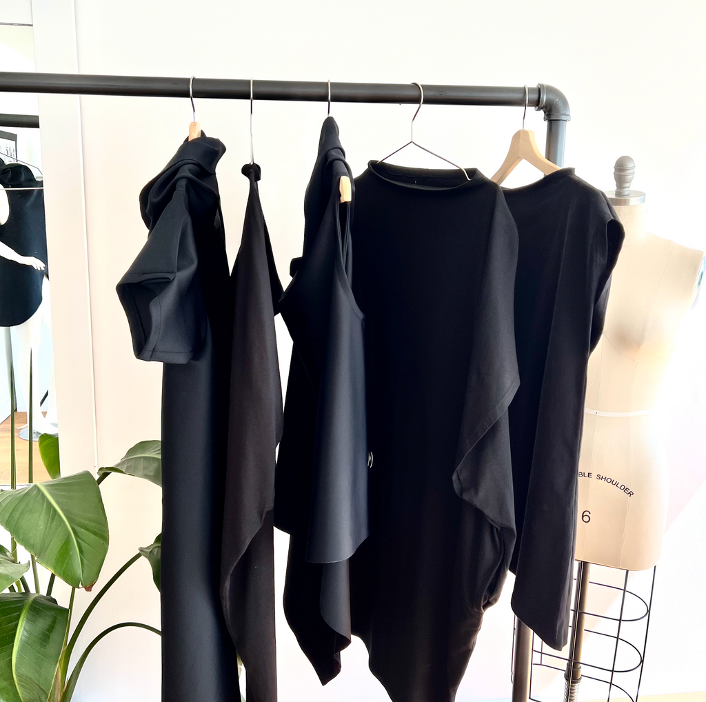 A rack of clothing with organic cotton black shift dress in knee length with pockets, long & asymmetrical cut by Malaika New York