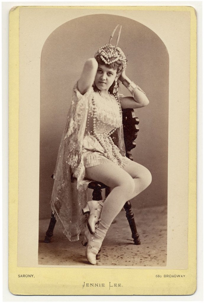 1890. Jennie Lee sitting on a chair,  holding a hair-dress on her head.