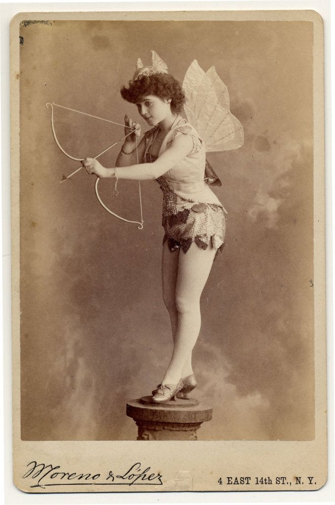 1890. Miss Murdock on pedestal, with fabric wings and horns shooting a bow and arrow.