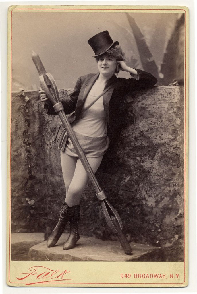 1890. Vernona Jabeau, in high boots, hat, holding a candle in a long holder.