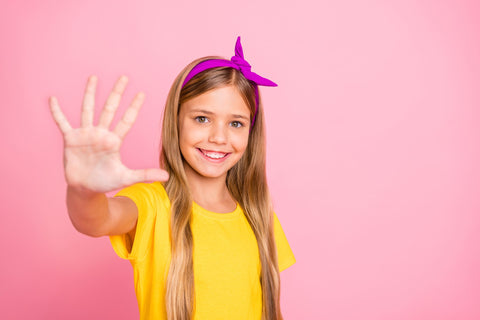 Girl with blond hair and yellow shirt smiling and holding five fingers up in front of a pink background.