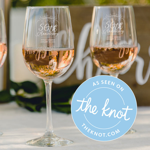 2 wine glasses are filled with rosé and sit atop a table wrapped in white linen. The glasses are etched with the words " 50th Anniversary. Anna & George. Established 1972."