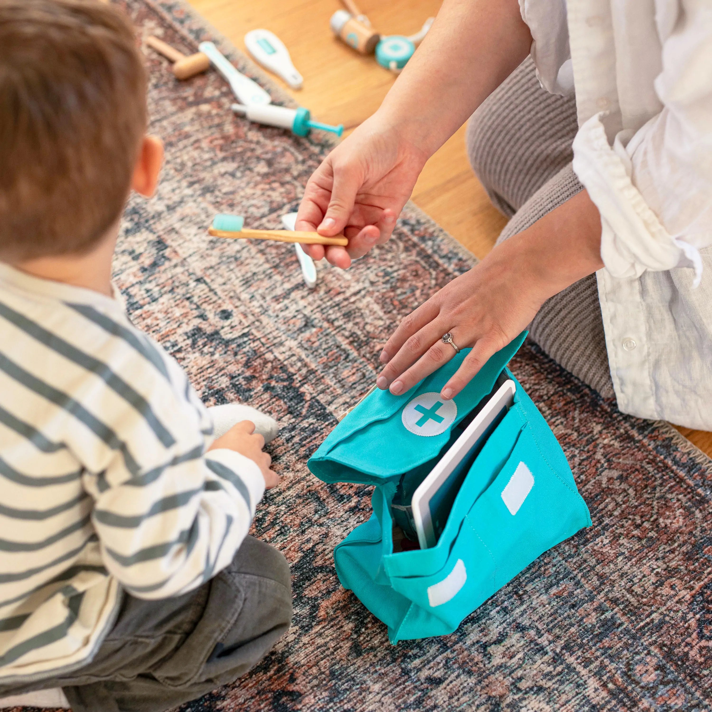 An adult and child play with the Tiny Land® Doctor Kit for Kids on a patterned rug. The adult holds a wooden thermometer as the open Tiny Land medical playset displays various pretend toys inside.