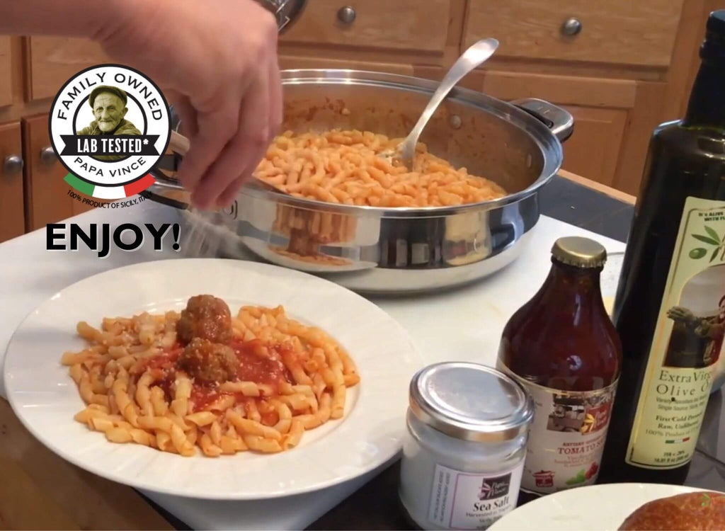 Hand serving a plate of Papa Vince Busiate pasta with meatballs, flanked by a jar of sea salt and bottles of tomato sauce and extra virgin olive oil, with a 'Family Owned, Lab Tested' seal and the word 'ENJOY!' displayed prominently.