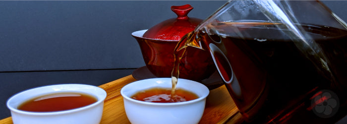 dark pu-erh tea is poured into two small white tasting cups in front of a red gaiwan