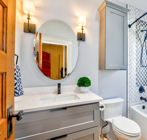 A modern bathroom with an oval mirror over the sink. A small green plant sits on the counter to the right of the sink, and a tall cabinet over the toilet provides storage space. The sink cabinet features two drawers with black edge pulls on a gray painted cabinets. Two square modern lights flank the mirrow. Off to the side is a shower with white subway tile and blue fixtures.