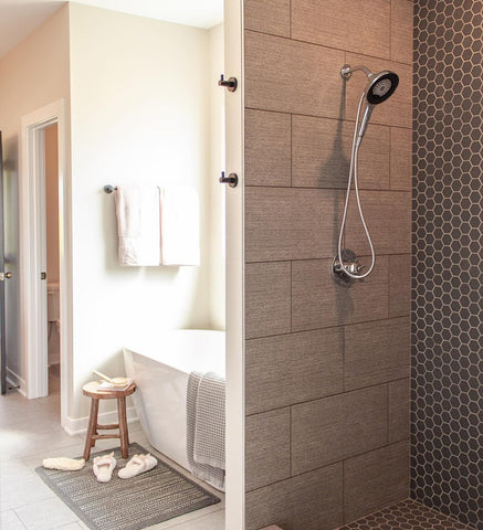 A modern shower with two robe hooks is in the foreground, while a deep soaking tub sits in an alcove behind the shower. Sunlight fills the space. 