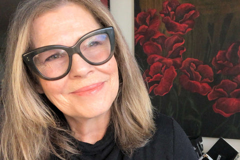 Conscious brand SAPPHO founder JoAnn Fowler smiling, head tilted wearing big glasses with an open professional makeup kit in the background