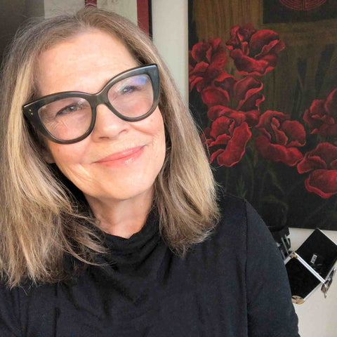 Makeup Artist and Founder JoAnn Fowler smiling with big glasses, sitting in front of a painting and an open makeup kit