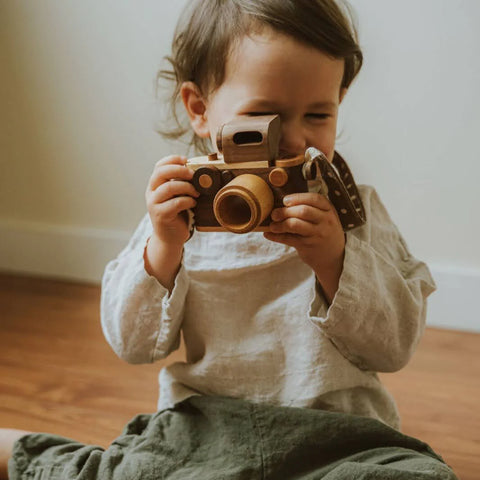 photo shows a toddler girl looking through a wooden toy camera pretending to take photos of mom.