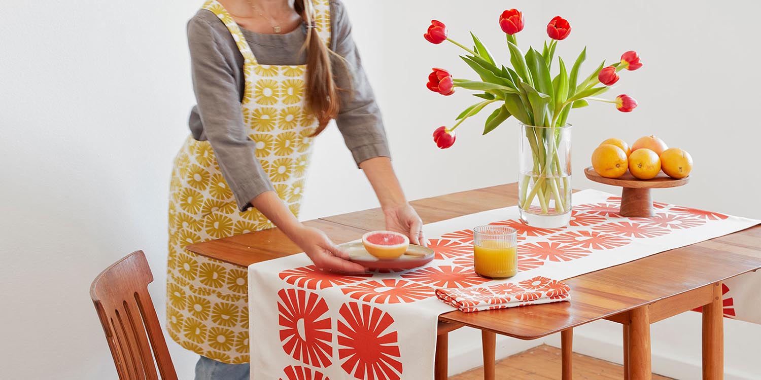 Skinny laMinx table runner, napkins and aprons styled on a tabletop. Skinny laMinx home décor products are available at Sarza home goods and furniture store in Rye New York.