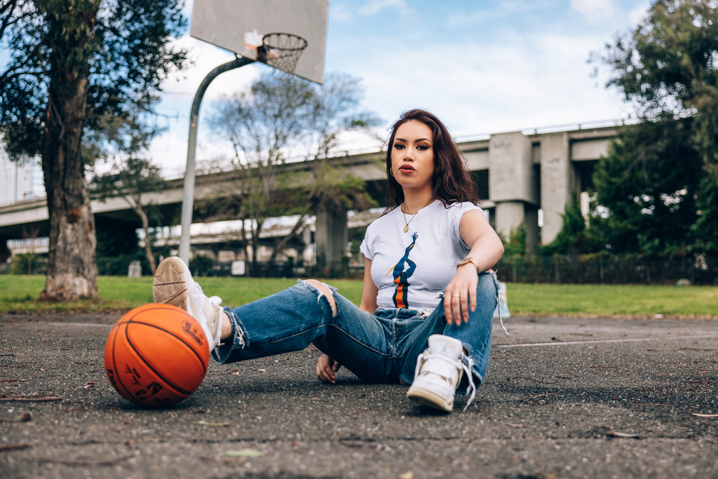 Girl model seated on asphalt with one leg on a basketball wearing white tee with Thunder rendition.