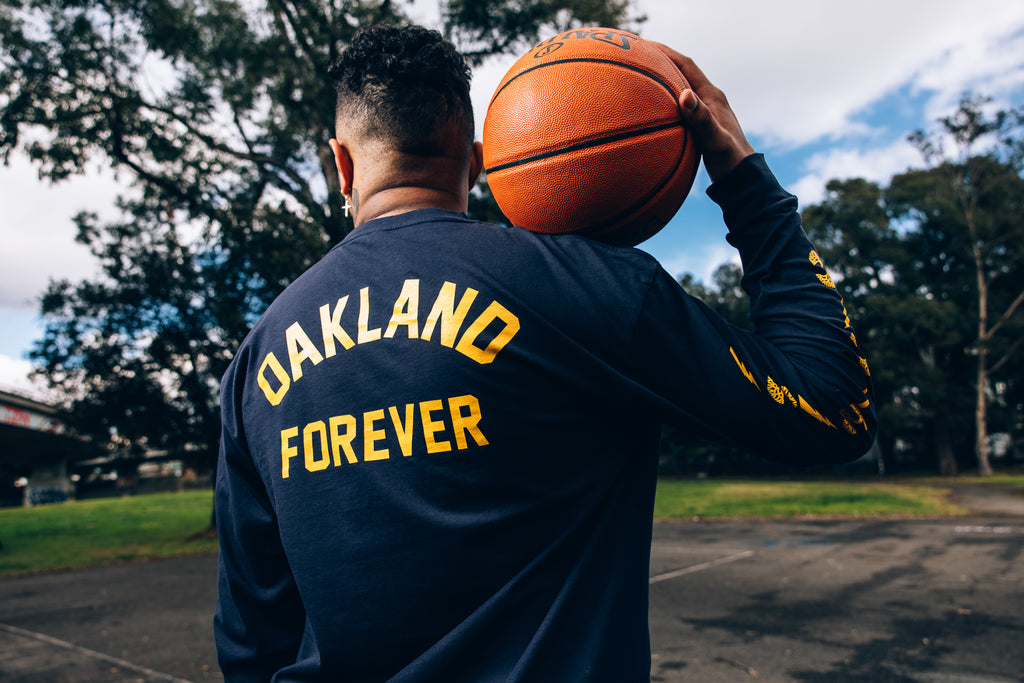 Model holding basketball on outdoor court, featuring Oakland Forever back print on navy long sleeved tee.