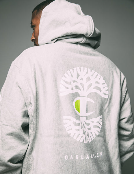 Model wearing grey pullover hoodie with white and green print of lockup of Champion C logo and Oaklandish tree logo.
