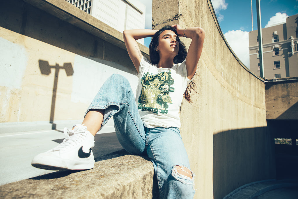 Girl model on rooftop in jeans and sneakers wearing a cream tee with Joshua mays design.