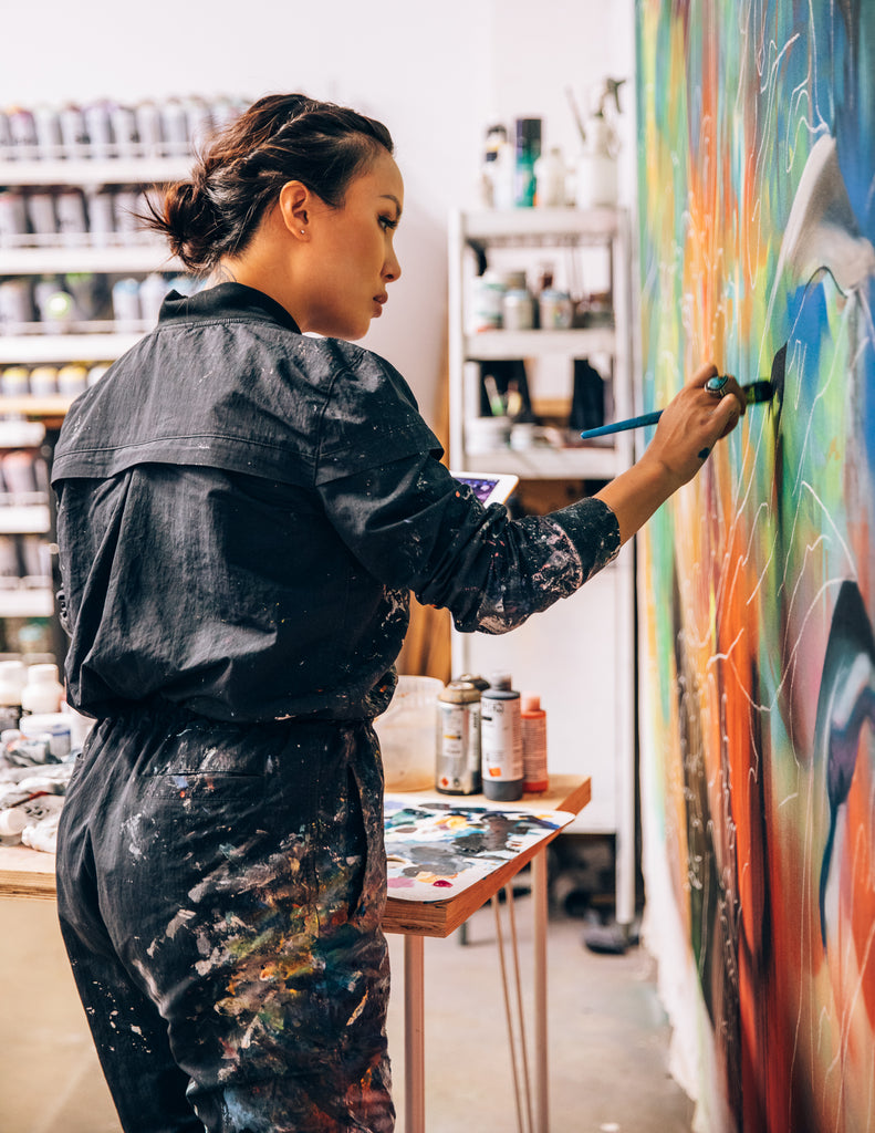 Hueman at work in her studio, wearing black jumpsuit splattered with paint.