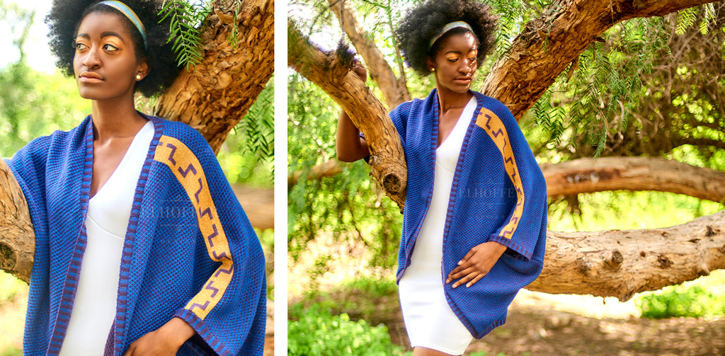 Armani (a tall and thin dark skinned model with black afro hair) models the Galactic Twilight Dolman over a white fitted dress. The dolman has an open front and is made from a blue knit with gold designs down the arms.