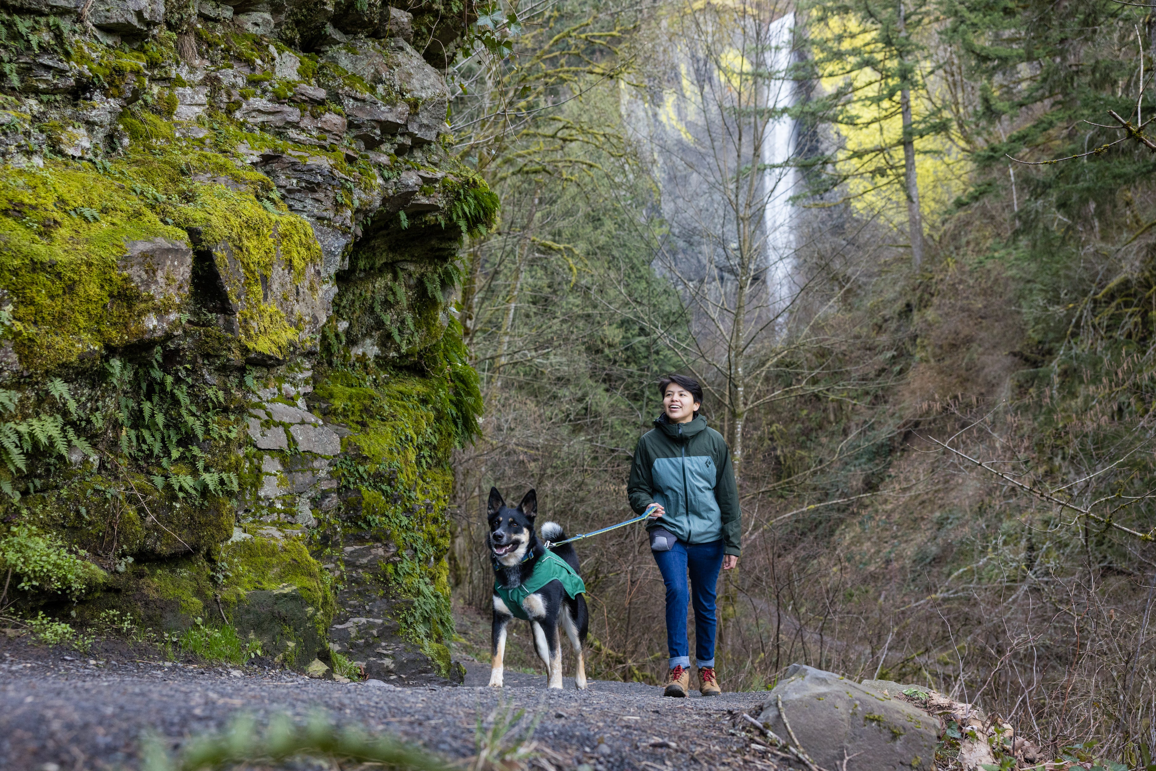 A woman and her dog walk on a trail by a waterfall. 