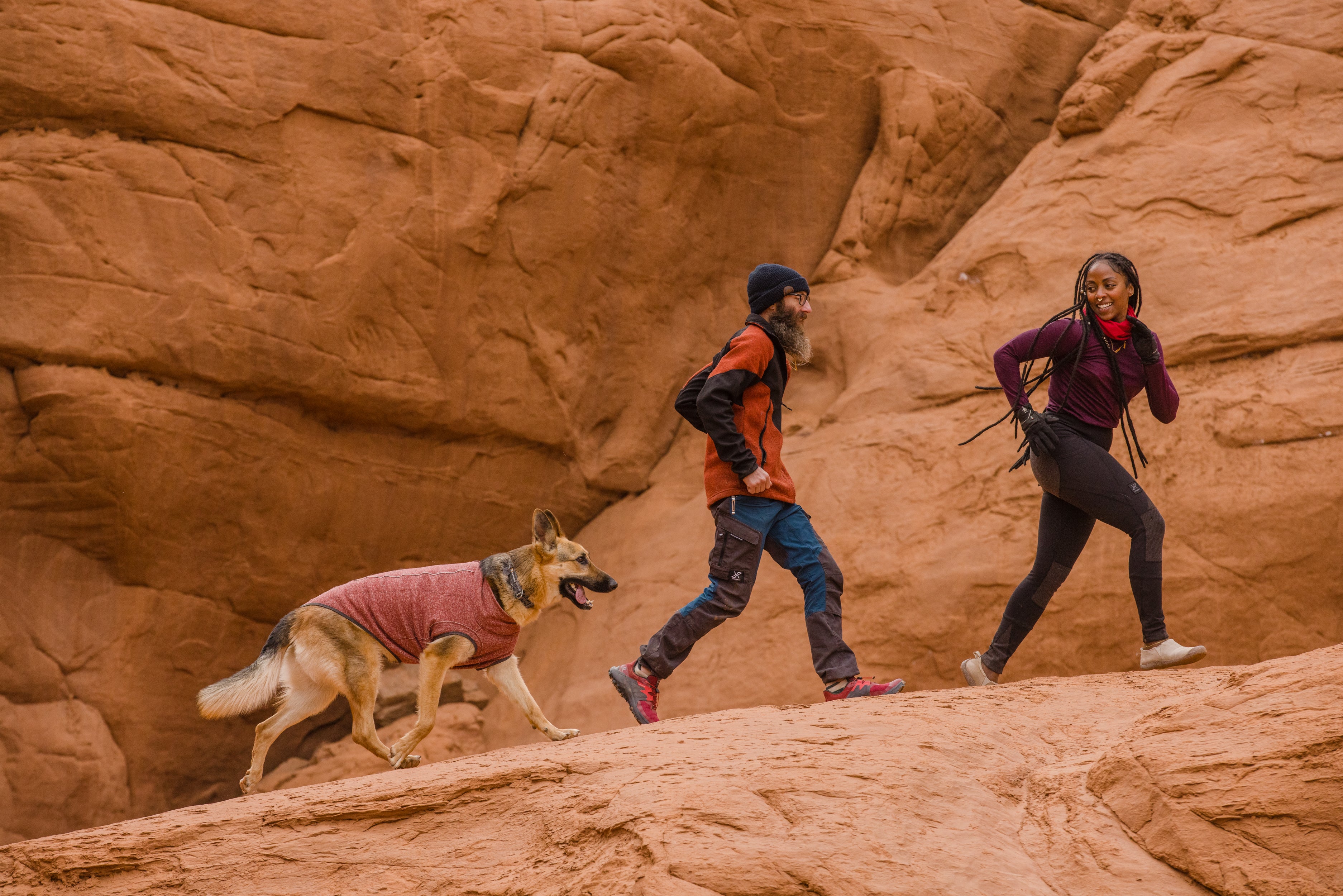 A woman and man run on a desert trail with their dog who is wearing a Hemp Hound™ Sweater. 