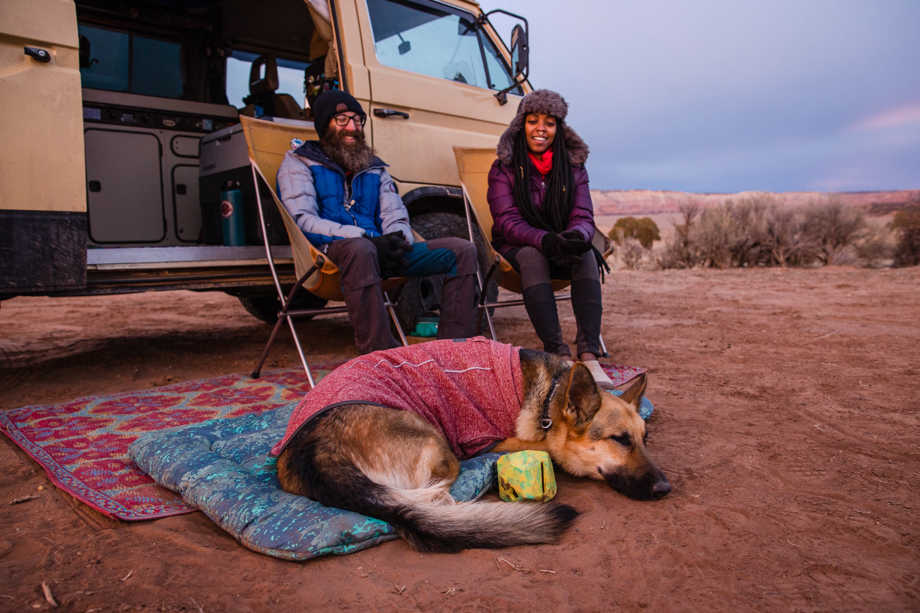 A woman and man sit outside their camper van while their dog, who is wearing a Hemp Hound™ Sweater, rests. 