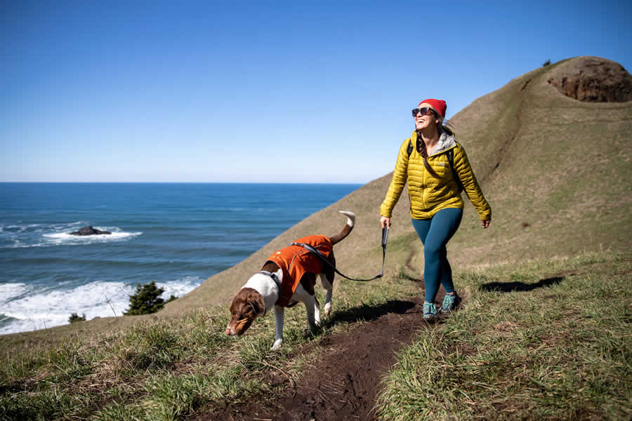 Woman hikes along Oregon coast in the sun with dog on leash clipped to overcoat fuse.