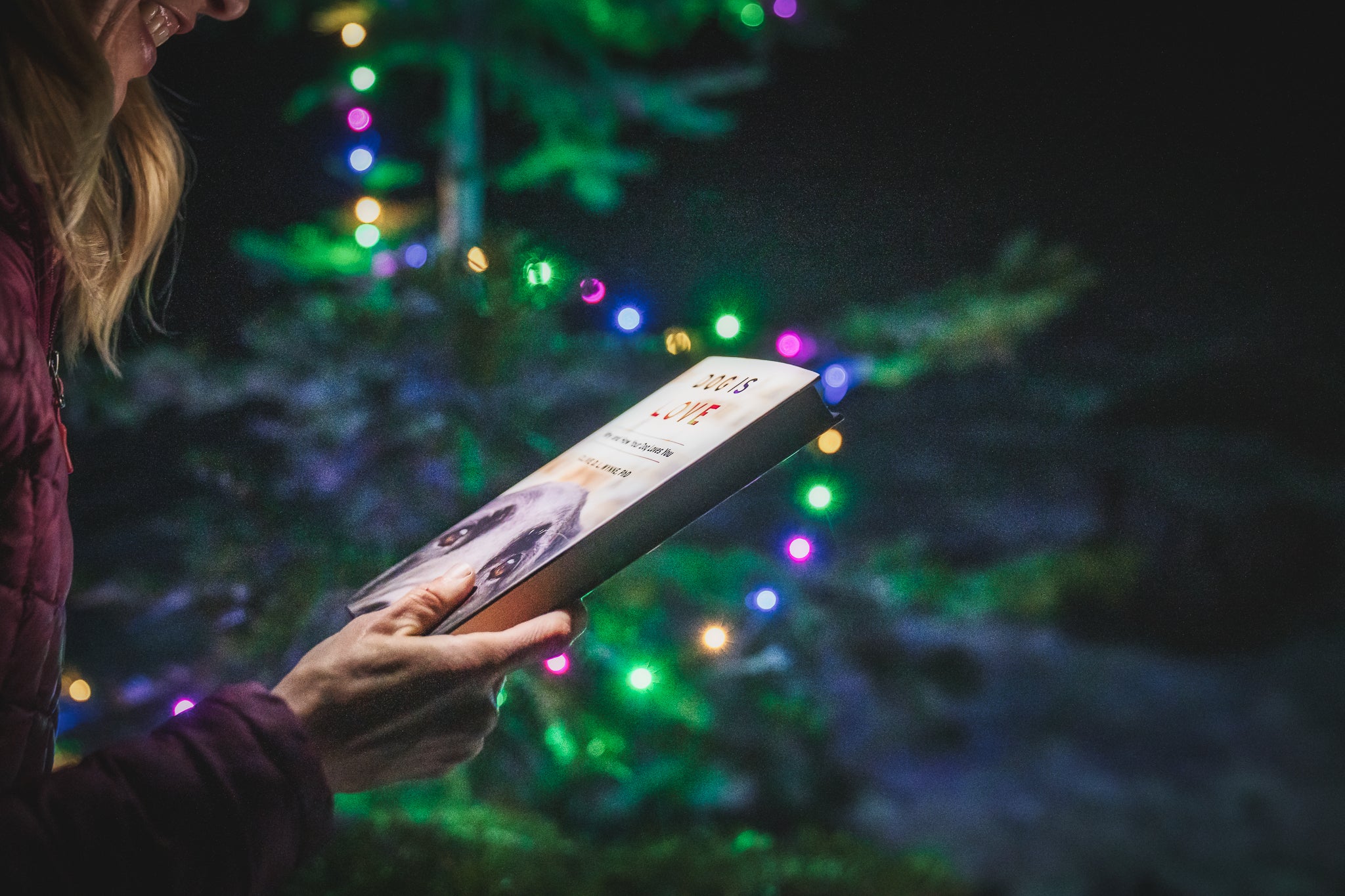 A woman holds a book by a Christmas tree. 