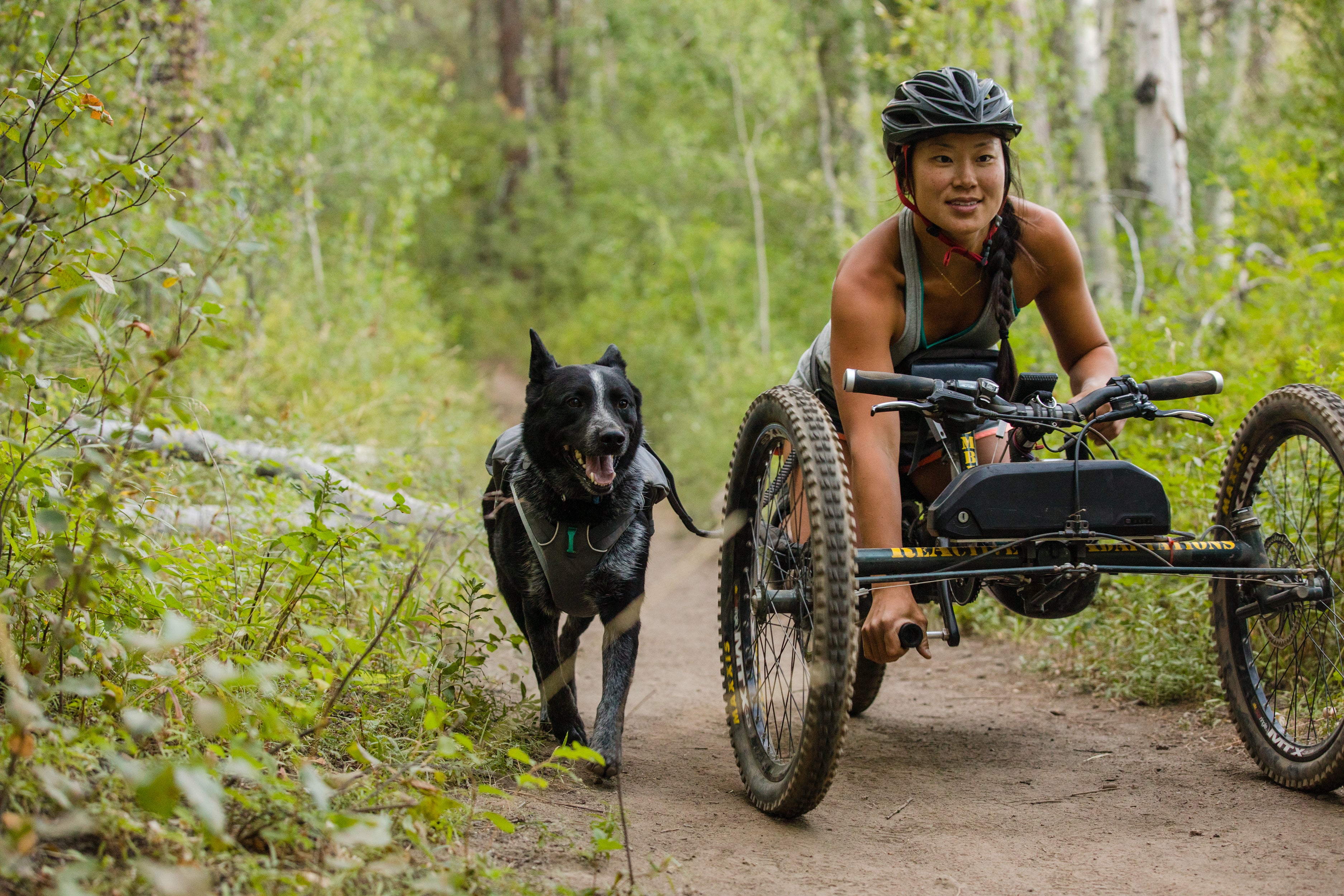 A woman rides a mountain bike that is accessible for her different physical abilities with her dog running beside her. 