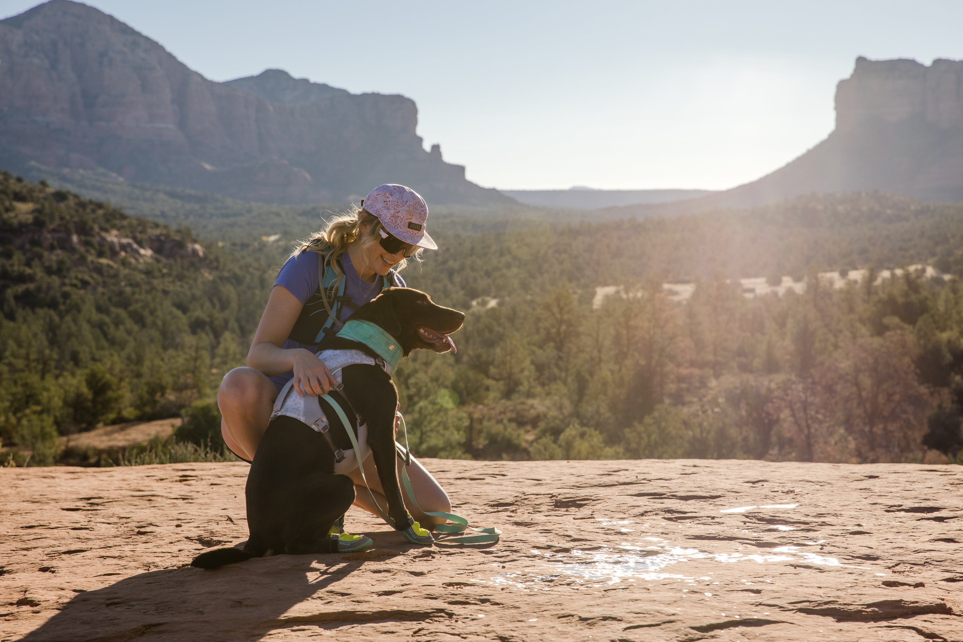 A woman and her dog sit and look at the view while hiking in the mountains. 