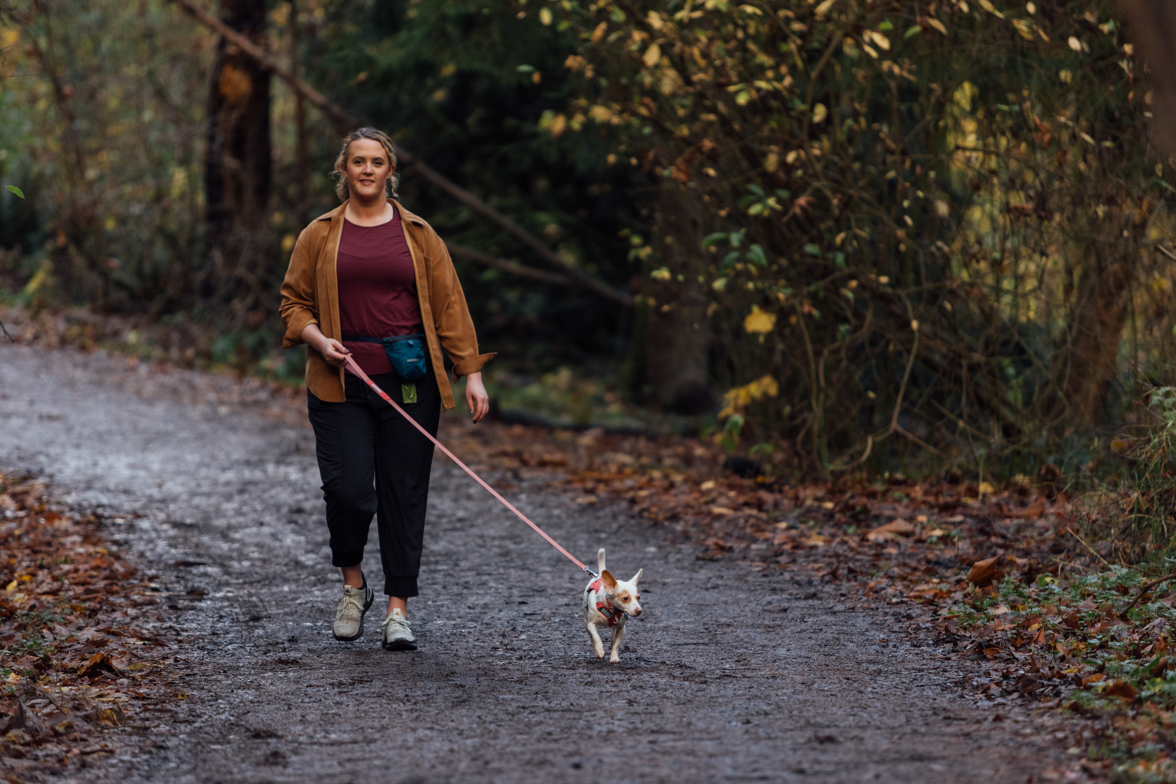 A woman walks with her dog on a trail in the woods.