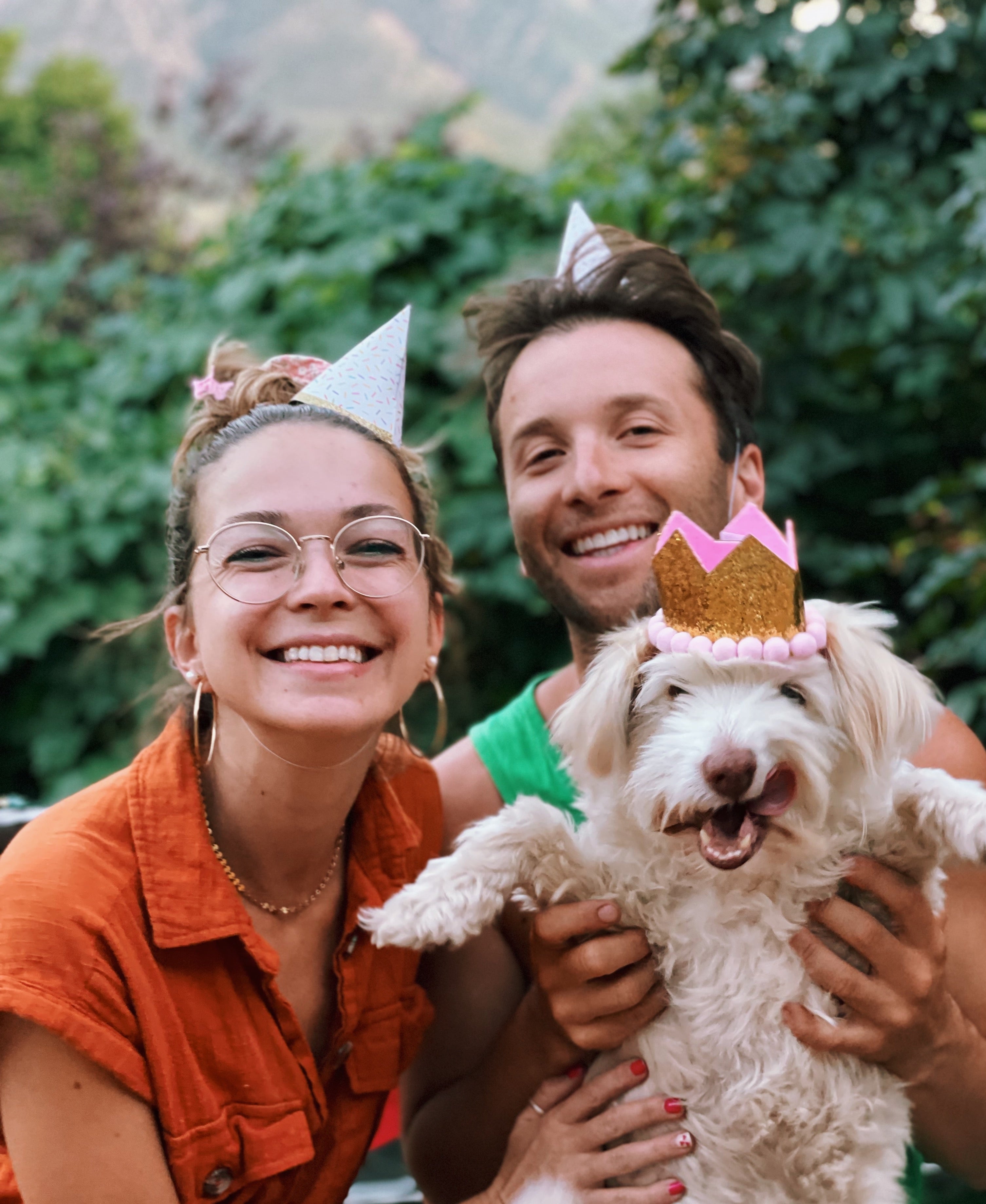A woman and man wearing birthday hats hold up their dog who is wearing a crown. 