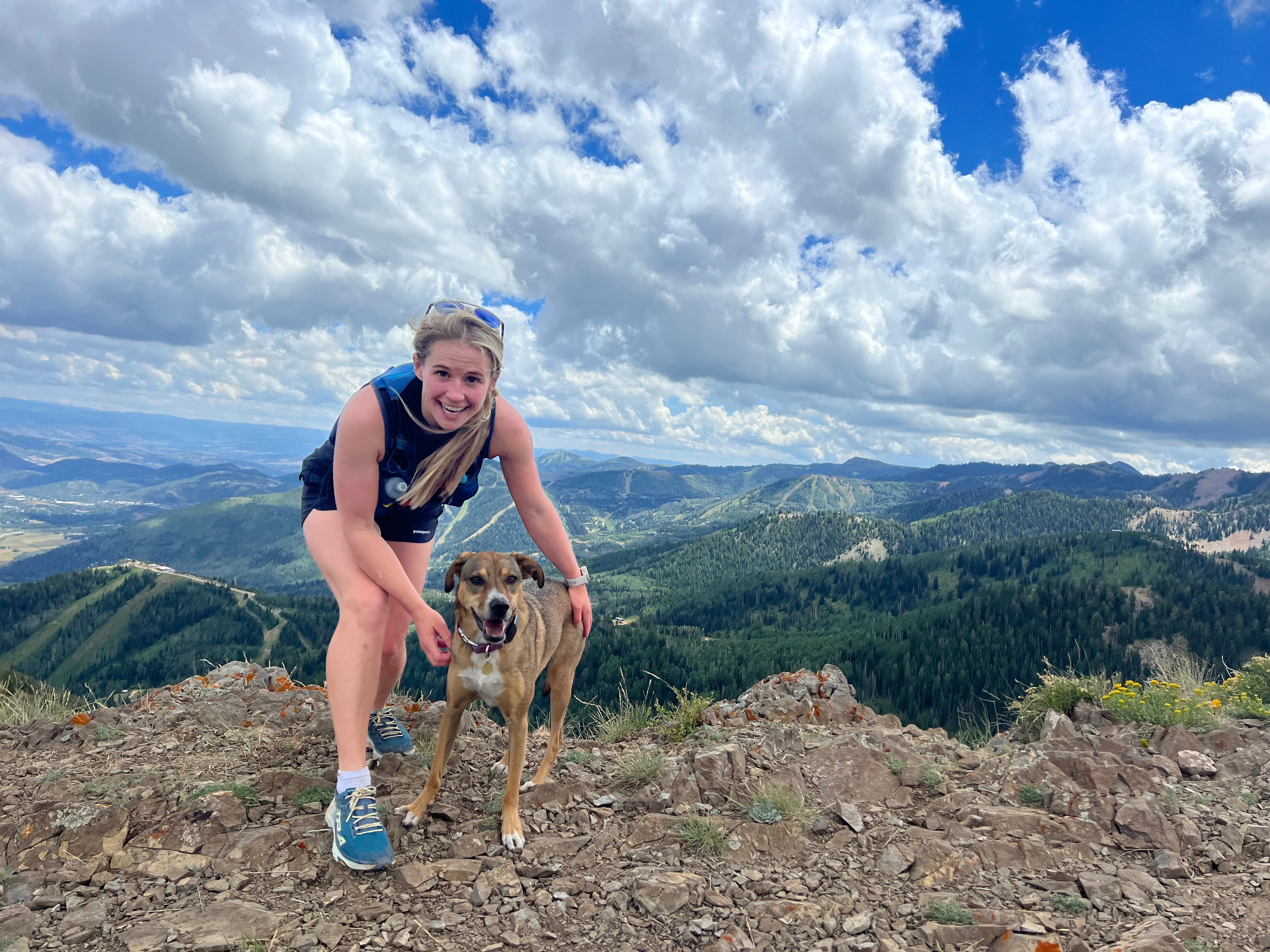 A woman poses with her dog on the top of a mountain. 