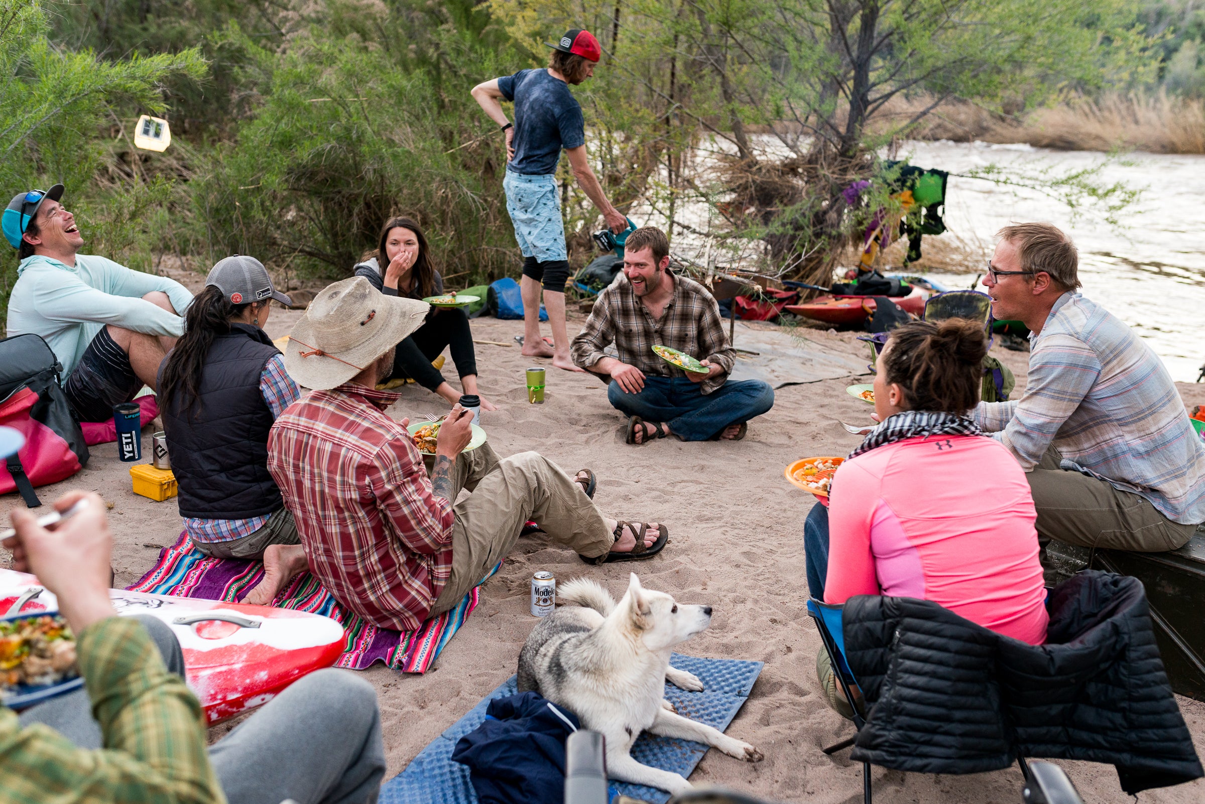 A group of people and a dog sit on the sand while camping and having a meal. 