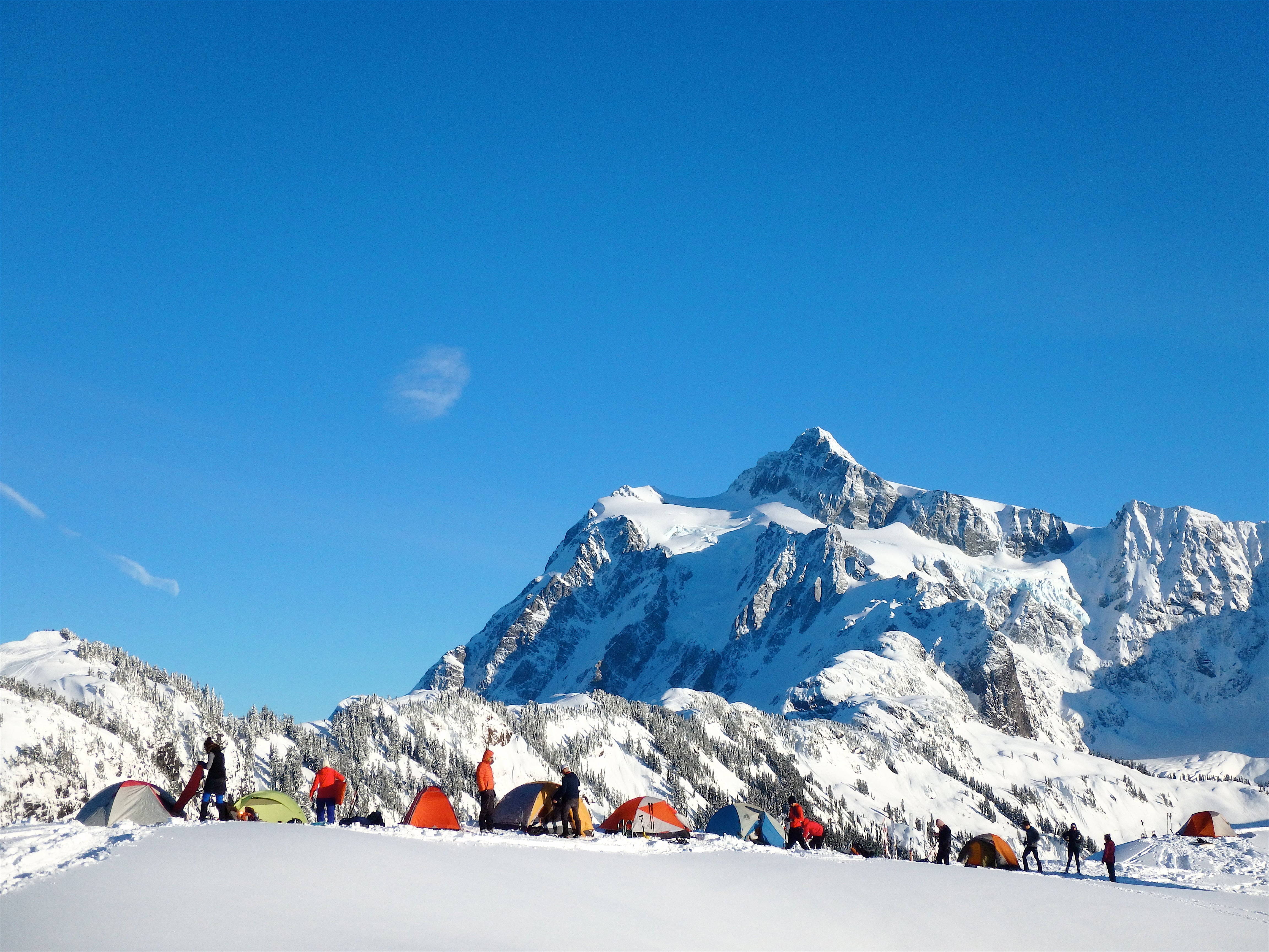 A group of people set up their tents in the snow, with mountains in the background. 