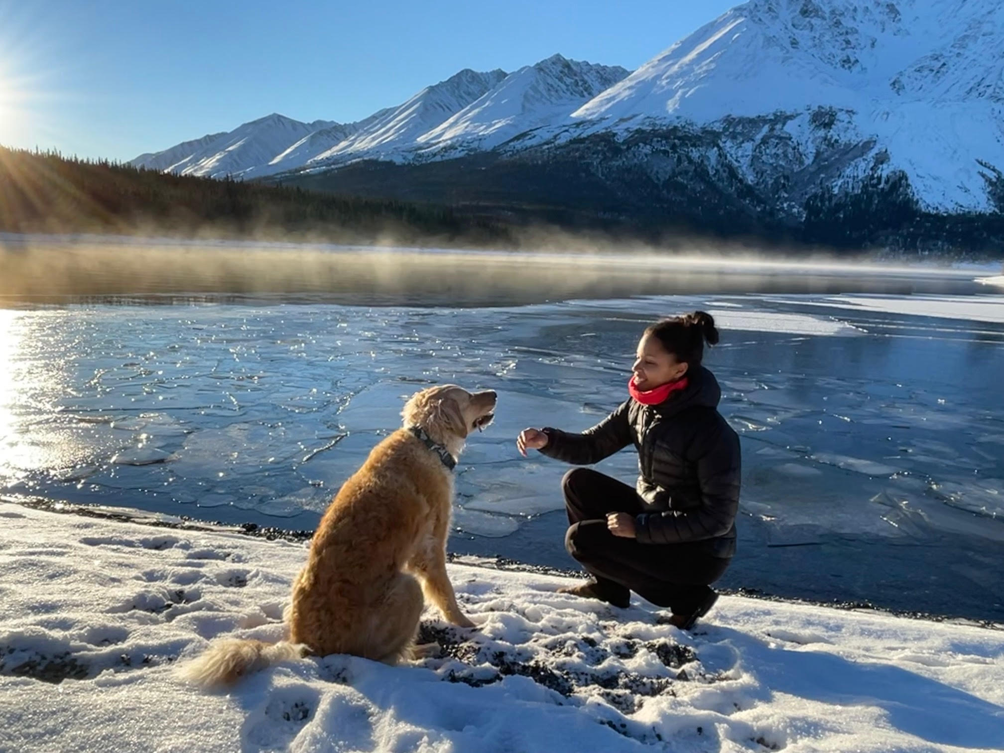 A woman crouches down by her dog in the snow by a lake in the mountains. 