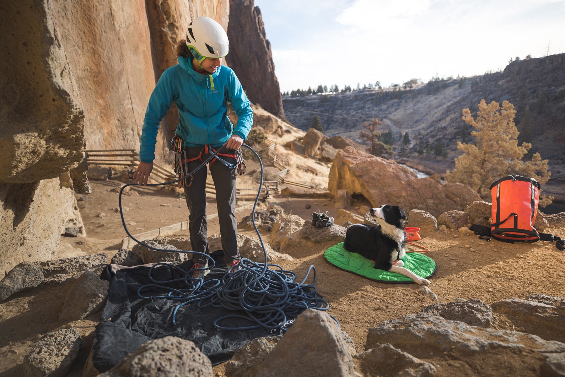 Woman flakes out climbing rope while dog lays on highland dog sleeping bag.