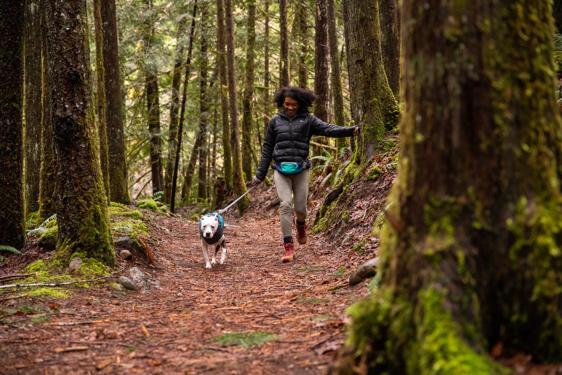 Woman and dog skip through green forested trail.