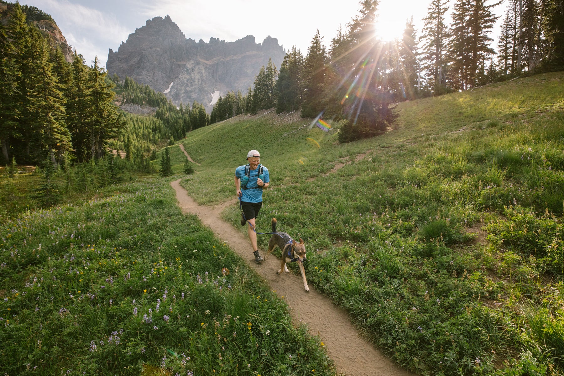 Dog and human run along trail with wildflowers.