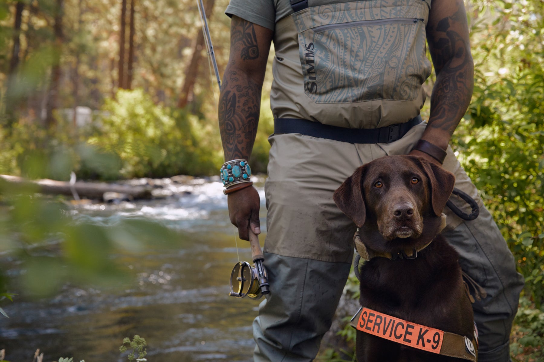 Chad in fly fishing gear and dog axe in service dog vest pose by the river close up.