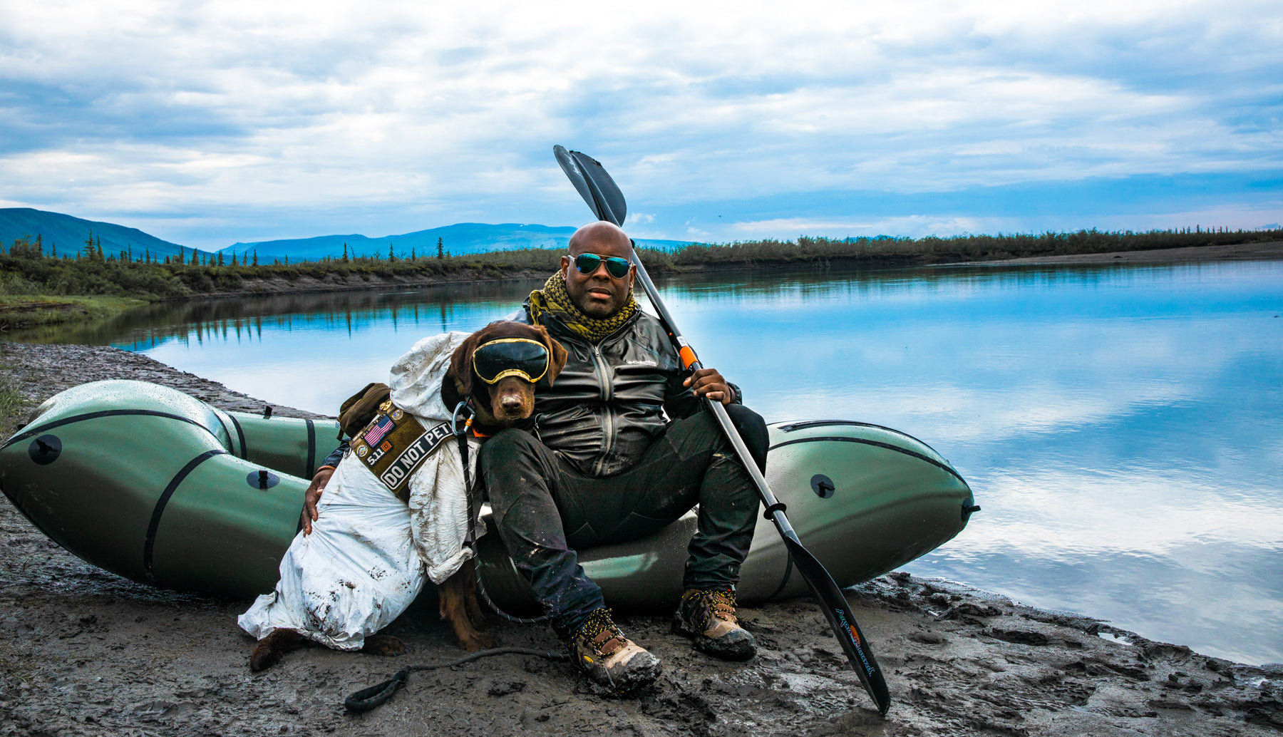 Chad and Axe sit by a packraft next to a lake with very blue skies in the background.