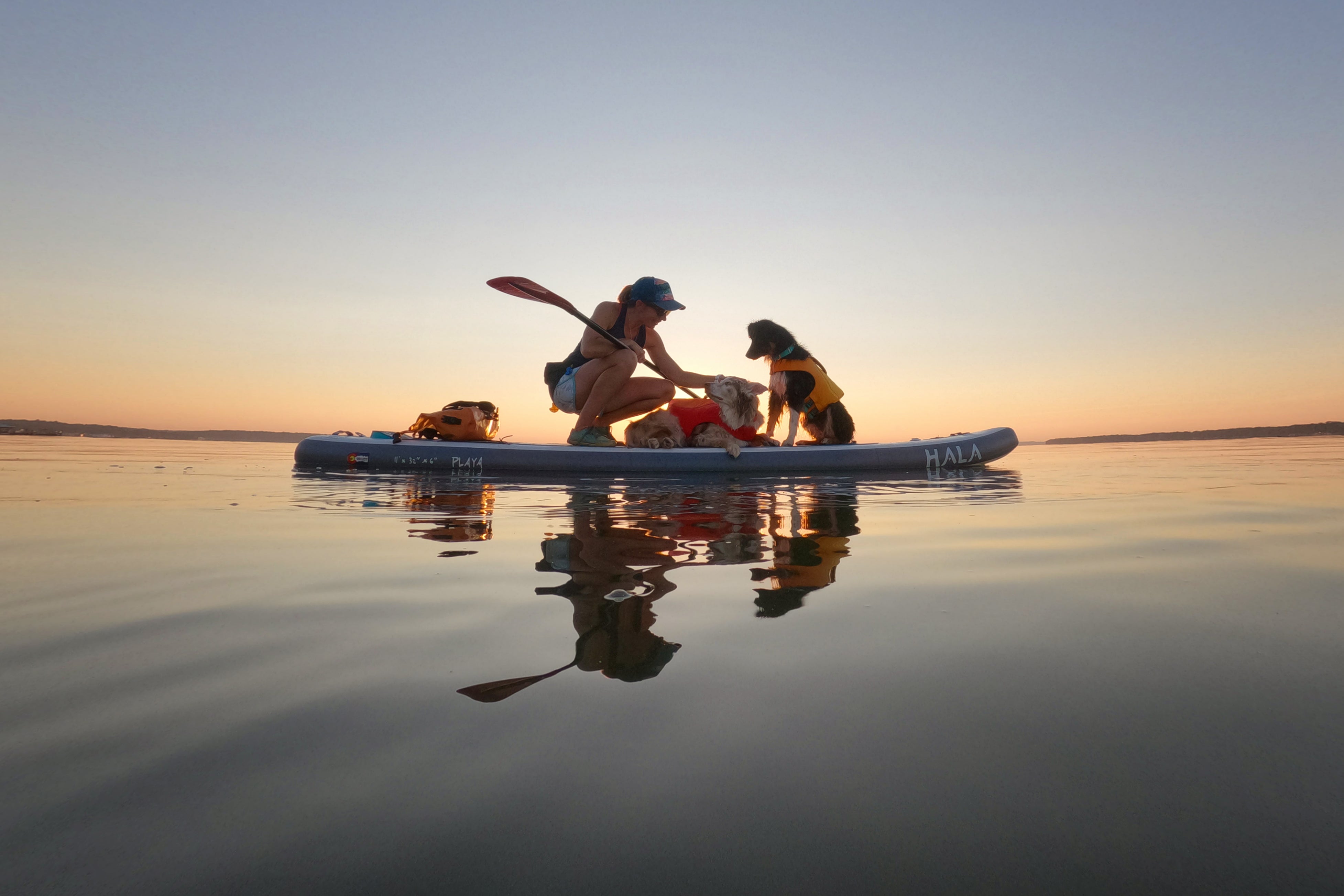 A woman kneels down on her paddleboard to pet her two dogs. 