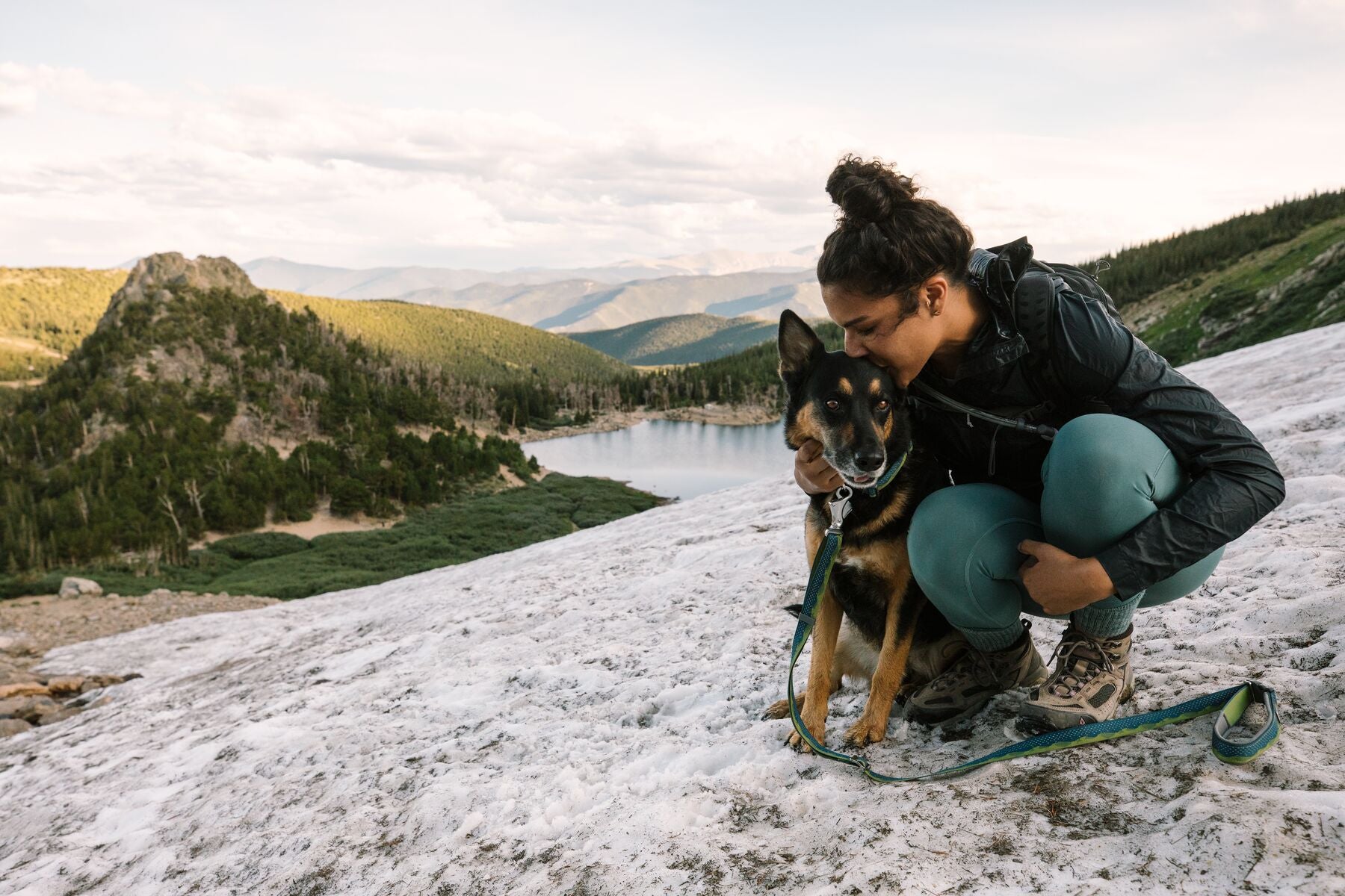 Woman gives dog wearing flat out collar and leash a kiss while sitting on the snow above an alpine lake.